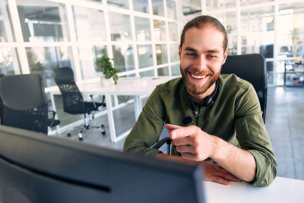 Technical support operator working with headset in office. Smiling handsome man working as call centre operator, speaking to customer. Happy businessman working remotely while doing video conference. - Photo, Image