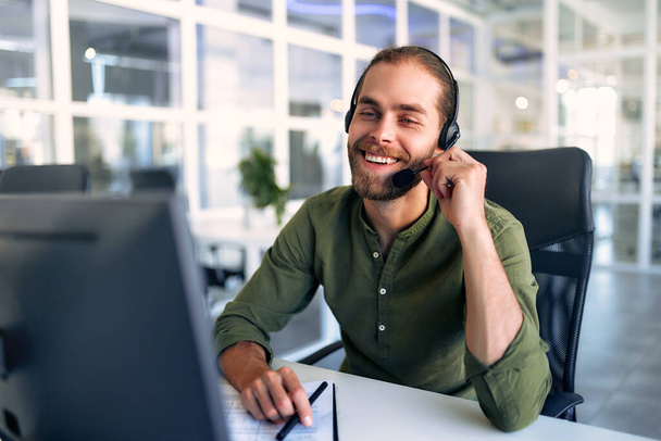 Διαχειριστής τεχνικής υποστήριξης που εργάζεται με ακουστικά στο γραφείο. Χαμογελώντας όμορφος άντρας που δουλεύει ως τηλεφωνητής, μιλώντας στον πελάτη. Ευτυχής επιχειρηματίας που εργάζονται εξ αποστάσεως, ενώ κάνει τηλεδιάσκεψη. - Φωτογραφία, εικόνα