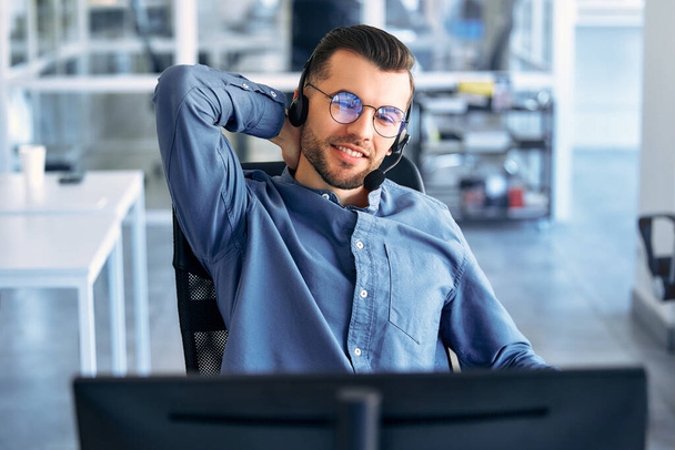 Technical support operator working with headset in office. Smiling handsome man working as call centre operator, speaking to customer. Happy businessman working remotely while doing video conference. - Photo, Image