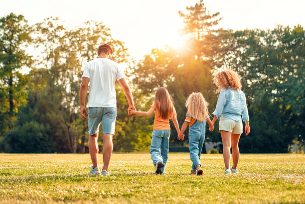 Szczęśliwy młody ojciec rodziny, mama i dwoje dzieci siostry spacerując, biegając i bawiąc się na łące w parku w ciepły słoneczny dzień, bawiąc się razem i ciesząc się przyrodą w dzień wolny. - Zdjęcie, obraz