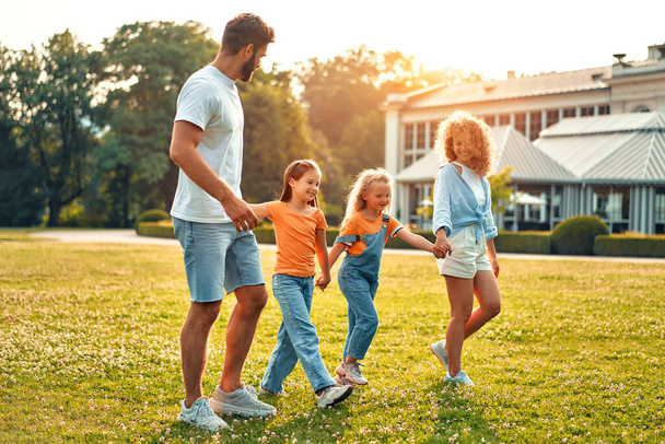 Ευτυχισμένη νεαρή οικογένεια μπαμπάς, μαμά και δύο αδελφές παιδιά με τα πόδια, τρέχει και παίζει στο λιβάδι στο πάρκο σε μια ζεστή ηλιόλουστη μέρα, διασκεδάζοντας μαζί και απολαμβάνοντας τη φύση σε μια μέρα μακριά. - Φωτογραφία, εικόνα
