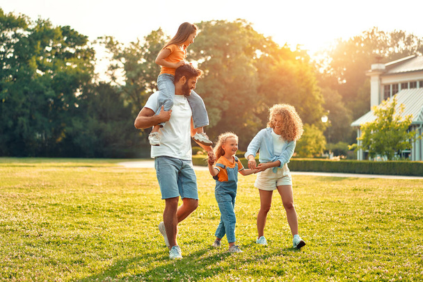Boldog fiatal család apa, anya és két gyermek testvér sétál, fut és játszik a réten a parkban egy meleg napsütéses napon, jól érzik magukat együtt, és élvezik a természet egy szabadnapon. - Fotó, kép