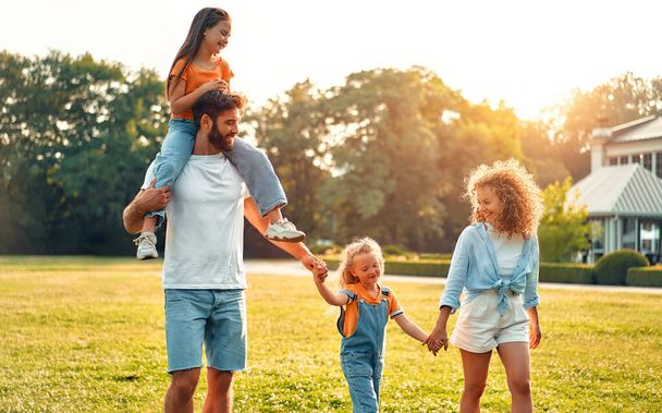 Glückliche junge Familie Vater, Mutter und zwei Kinder Schwestern gehen, laufen und spielen auf der Wiese im Park an einem warmen, sonnigen Tag, haben Spaß zusammen und genießen die Natur an einem freien Tag. - Foto, Bild