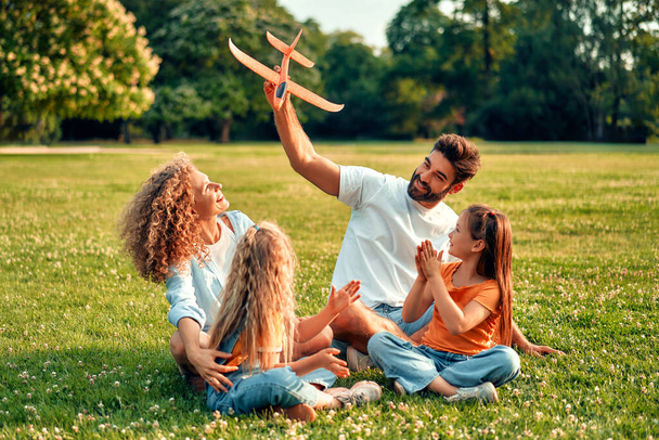 Szczęśliwy młody ojciec rodziny, mama i dwie córki bawiące się samolotem siedzącym na łące na trawie w parku w ciepły słoneczny dzień, bawiące się w wolny dzień. - Zdjęcie, obraz