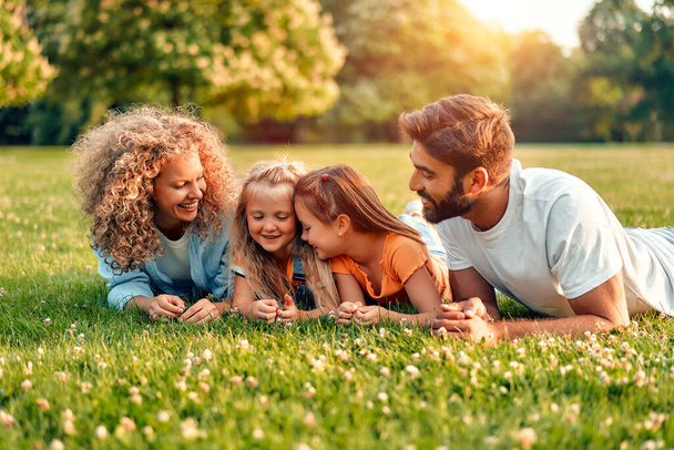 Szczęśliwy młody ojciec rodziny, mama i dwie córki leżące na trawie na łące w parku w ciepły słoneczny dzień, bawiące się razem i relaksujące w dzień wolny. - Zdjęcie, obraz