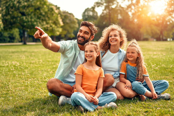 Feliz padre de familia joven, mamá y dos hijas sentadas en la hierba en un prado en el parque en un día cálido y soleado, divirtiéndose y trenzando juntos, relajándose en un día libre. - Foto, imagen