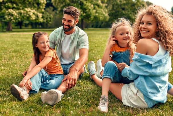 Χαρούμενη νεαρή οικογένεια μπαμπάς, μαμά και δύο κόρες κάθονται στο γρασίδι σε ένα λιβάδι στο πάρκο σε μια ζεστή ηλιόλουστη μέρα, διασκεδάζοντας και πλένοντας μαζί, χαλαρώνοντας σε μια μέρα μακριά. - Φωτογραφία, εικόνα