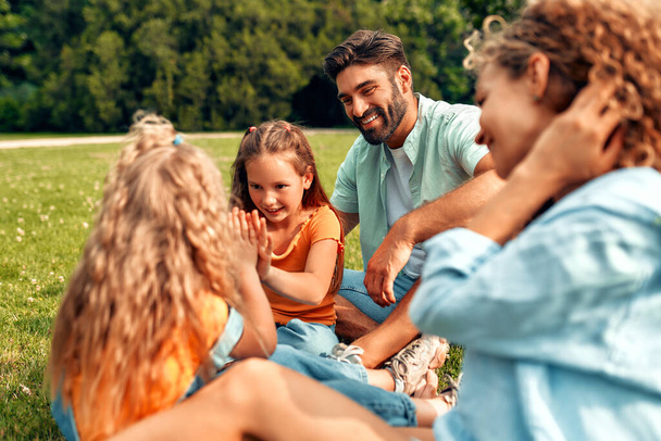 Feliz padre de familia joven, mamá y dos hijas sentadas en la hierba en un prado en el parque en un día cálido y soleado, divirtiéndose y trenzando juntos, relajándose en un día libre. - Foto, imagen