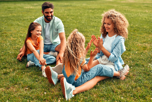 Счастливая молодая семья папа, мама и две дочери сидят на траве в лугу в парке в теплый солнечный день, весело и plaing вместе, расслабляясь в выходной день. - Фото, изображение