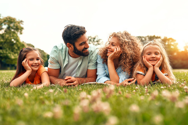 Onnellinen nuori perhe isä, äiti ja kaksi tytärtä makaa nurmikolla niityllä puistossa lämpimänä aurinkoisena päivänä, hauskaa yhdessä ja rentouttavaa vapaapäivänä.. - Valokuva, kuva
