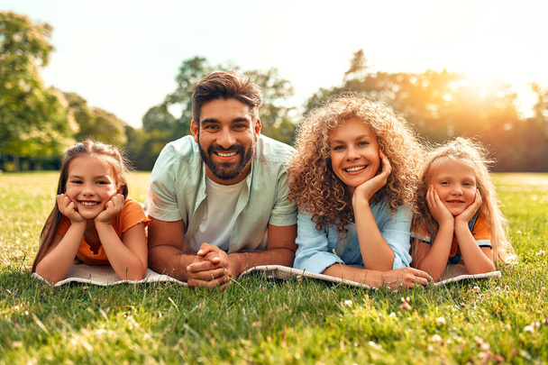 Feliz pai de família jovem, mãe e duas filhas deitadas na grama em um prado no parque em um dia ensolarado quente, se divertindo juntos e relaxando em um dia de folga. - Foto, Imagem