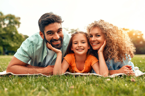 Mutlu, genç bir aile babası, anne ve kızı güneşli bir günde parkta çimlerin üzerinde uzanıyor, birlikte eğleniyor ve tatil günlerinde dinleniyorlar.. - Fotoğraf, Görsel