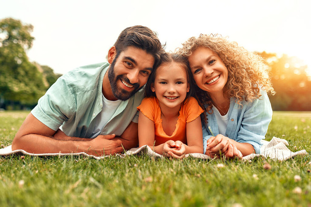 Szczęśliwy młody ojciec rodziny, mama i córka leżące na trawie na łące w parku w ciepły słoneczny dzień, bawiąc się razem i relaksując się w dzień wolny. - Zdjęcie, obraz