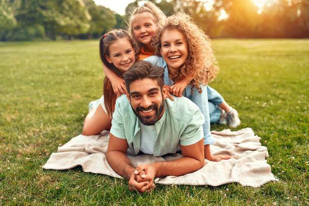 Šťastný mladý rodinný táta, máma a dvě dcery ležící na trávě na louce v parku za teplého slunečného dne, baví se spolu a relaxují na den volna. - Fotografie, Obrázek