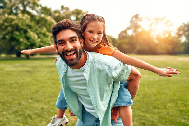 Boldog fiatal apa játszik a kislányával, háton fordítja, fut és szórakozik a parkban a réten egy meleg, napsütéses napon egy hétvégén. - Fotó, kép