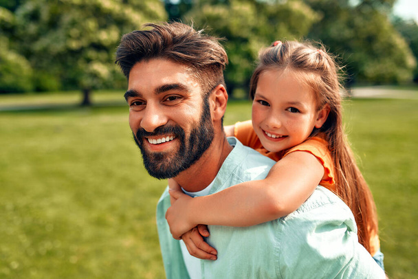 Szczęśliwy młody ojciec bawi się ze swoją córeczką, obracając ją na plecach, biegając i bawiąc się na łące w parku w ciepły słoneczny dzień w weekend. - Zdjęcie, obraz