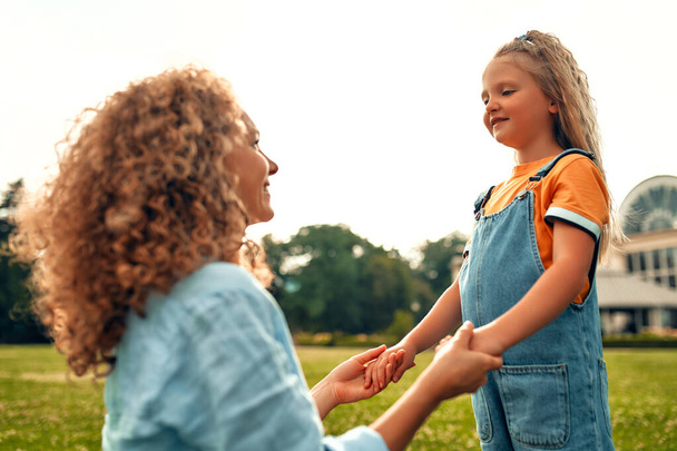 Прекрасная счастливая молодая мама держит за руки свою любимую маленькую дочь на лугу в парке в теплый солнечный день, играя и веселясь вместе. - Фото, изображение