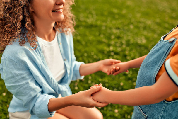Hermosa madre joven feliz sosteniendo las manos de su querida hija pequeña en el prado en el parque en un día cálido y soleado, jugando y divirtiéndose juntos. - Foto, Imagen