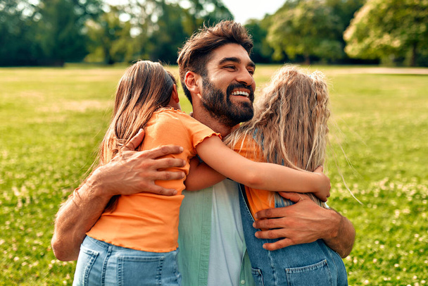 Glücklicher junger Vater umarmt seine beiden geliebten süßen Töchter, während er auf einer Wiese im Park sitzt, spielt und Spaß an einem warmen, sonnigen Tag hat. - Foto, Bild