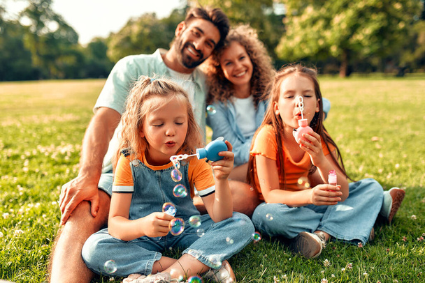 Szczęśliwy młody ojciec rodziny, mama i dwie córki dmuchanie bańki mydlane siedzi na łące na trawie w parku, bawiąc się i bawiąc się razem w ciepły słoneczny dzień wolny. - Zdjęcie, obraz