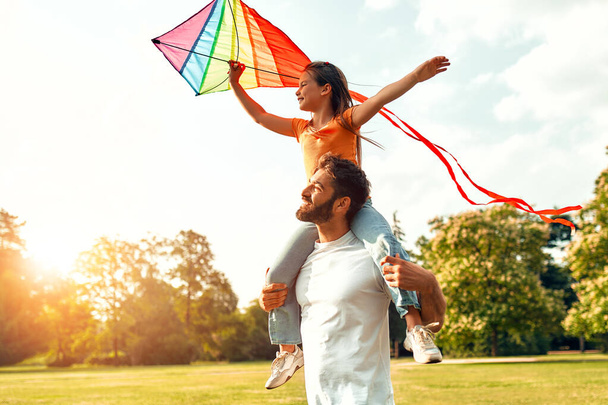 Щасливий молодий батько, граючи зі своєю коханою дочкою з повітрям барвистий змій на лузі в парку, розважається разом у теплі сонячні вихідні. - Фото, зображення