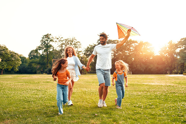 Χαρούμενη νεαρή οικογένεια μπαμπάς, μαμά και δύο κόρες τρέχει και παίζει με ένα πολύχρωμο χαρταετό σε ένα λιβάδι στο πάρκο, έχοντας τη διασκέδαση σε ένα ζεστό ηλιόλουστο Σαββατοκύριακο. - Φωτογραφία, εικόνα