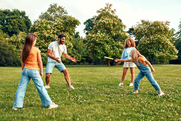 Szczęśliwy młody ojciec rodziny, mama i dwie córki bawiące się frisbee na łące w parku, bawiąc się razem w ciepły słoneczny dzień wolny. - Zdjęcie, obraz