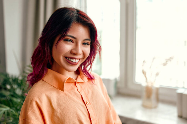 リビングルームやオフィスの自宅の窓に立ったシャツにピンク色の髪のストリークを持つ若い明るいアジアの女性のクローズアップの肖像画. - 写真・画像