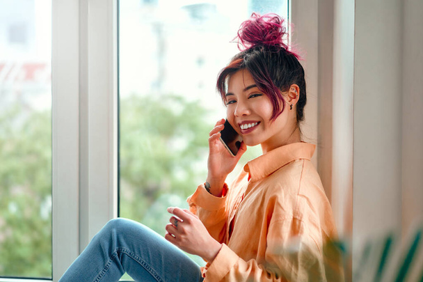 Giovane donna asiatica con lucenti ciocche di capelli rosa che parlano al telefono mentre si siede sul davanzale della finestra in soggiorno a casa. - Foto, immagini