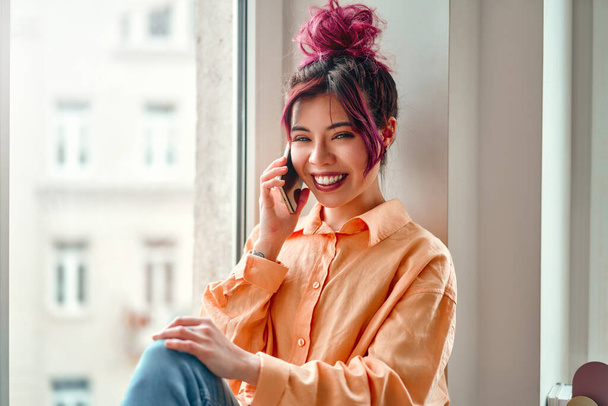 Młoda Azjatka z jasnoróżowymi włosami rozmawiająca przez telefon siedząc na parapecie przy oknie w salonie w domu. - Zdjęcie, obraz