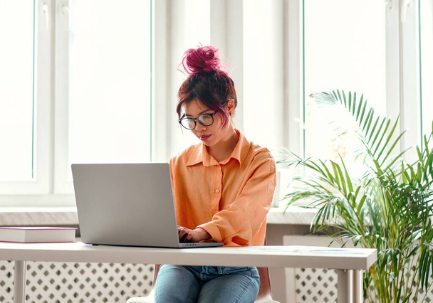Asiática joven con hebras de color rosa brillante de pelo sentado en una mesa con un ordenador portátil en casa en la sala de estar, trabajando o haciendo aprendizaje a distancia. - Foto, imagen