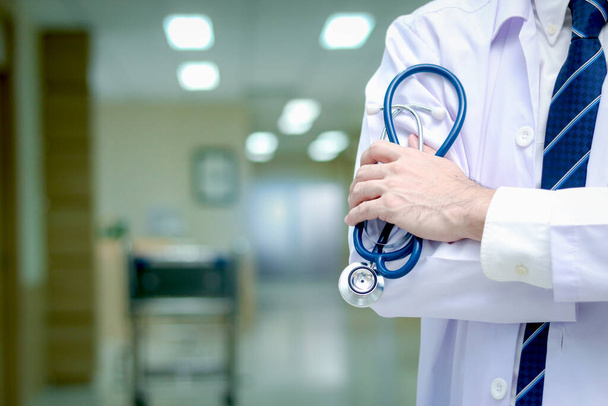 Pewny siebie lekarz w białej odzieży roboczej trzymający stetoskop stojący z rękami skrzyżowanymi w korytarzu kliniki medycznej, lekarz pracujący w szpitalu. koncepcja opieki medycznej i zdrowotnej. - Zdjęcie, obraz