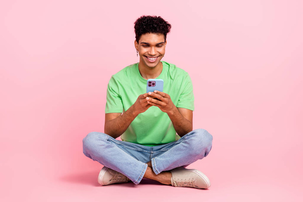 Full size photo of cheerful guy dressed green t-shirt sitting on floor look at smartphone read email isolated on pink color background. - Φωτογραφία, εικόνα