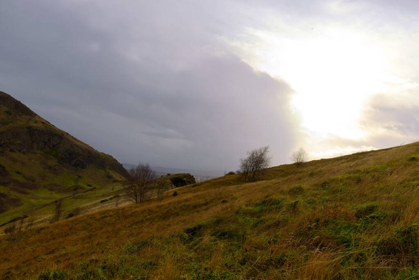 Experimente o drama celestial se desenrolar sobre o icônico Arthur Seat de Edimburgo. Esta fotografia capta o cume rochoso silhueta contra um pano de fundo de tons deslumbrantes do pôr do sol e nuvens expressivas - Foto, Imagem