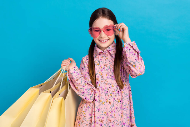 Photo portrait of pretty schoolkid hold shopping bags sunglass dressed stylish pink flower print outfit isolated on blue color background. - Foto, afbeelding