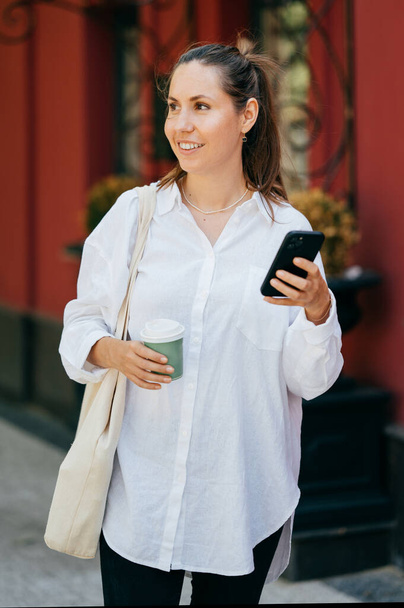 Fröhliche junge Frau genießt Kaffee, während sie mit ihrer Öko-Einkaufstasche spazieren geht. Hintergrund Rotes Haus. - Foto, Bild