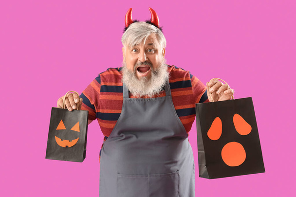 Шокированный зрелый мужчина, одетый на Хэллоуин с подарочными пакетами на розовом фоне - Фото, изображение