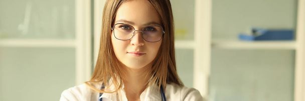Νεαρή όμορφη γυναίκα γιατρός με γυαλιά με στηθοσκόπιο κοιτάζοντας την κάμερα. Εκπαιδευόμενη που εργάζεται σε ιατρική κλινική, έννοια της ιατρικής εκπαίδευσης. - Φωτογραφία, εικόνα