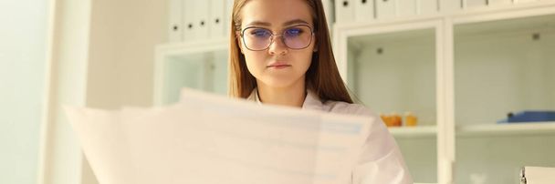 Nuori silmälasipäinen naislääkäri tekee paperitöitä klinikalla. Sairaanhoitaja työskentelee lääketieteellisten lomakkeiden kanssa istuessaan sairaalassa pöydässä. - Valokuva, kuva
