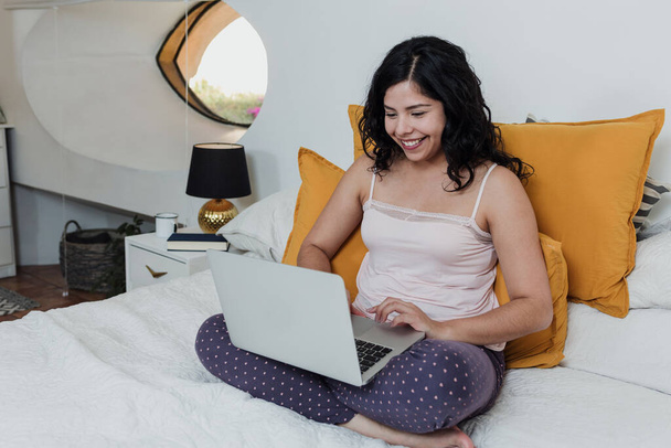 латинская молодая женщина сидит на кровати с ноутбуком дома, латиноамериканка в работе из дома или фрилансер и стиль жизни - Фото, изображение