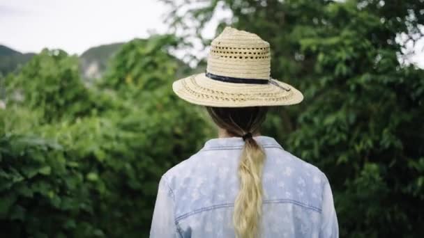 Rückansicht einer nicht wiederzuerkennenden blonden Bäuerin mit Strohhut, die auf einer üppigen Plantage spaziert - Filmmaterial, Video