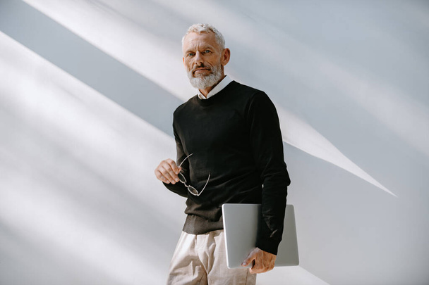 Portrait of confident senior man in smart casual wear holding laptop while standing against a wall - Foto, Bild
