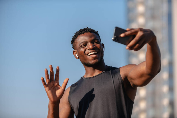 Happy young African athlete using smart phone and smiling while having video call outdoors - 写真・画像