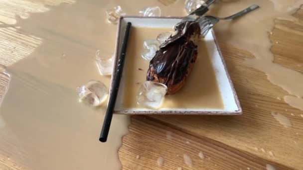 un vaso se rompió en una mesa de madera, capuchino café con leche derramado, trozos de vidrio en un plato con un pastel de macarrones y una paja que yace cerca, una molestia derramando café. limpieza en un restaurante - Imágenes, Vídeo