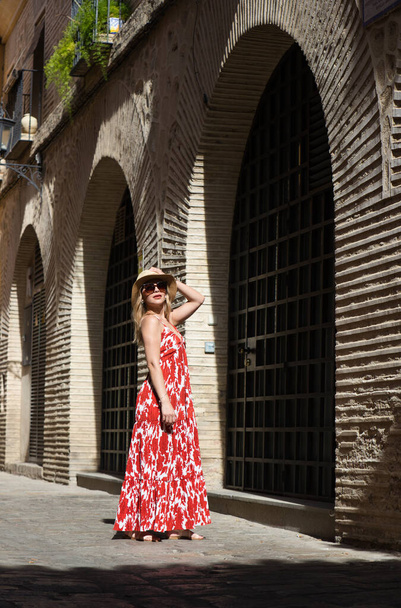 Mladá a krásná blondýnka se slunečními brýlemi a slaměným kloboukem na hlavě kráčí historickým centrem Seville. Žena je na dovolené a užívá si města. Ta žena je šťastná. - Fotografie, Obrázek