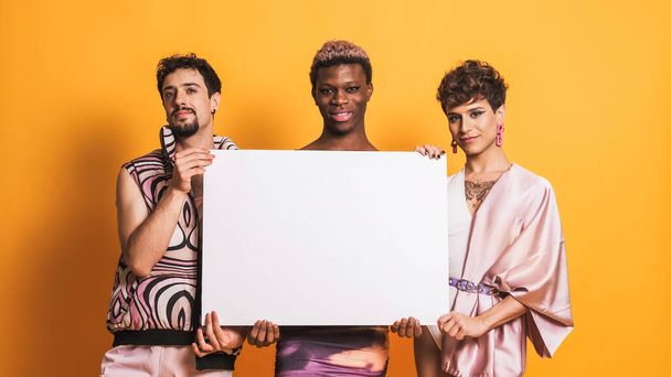 three multiethnic transsexual men holding together a white empty sign in a yellow background - Photo, Image