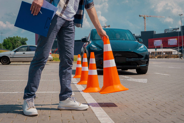Άντρας δάσκαλος οδήγησης στέκεται με πορτοκαλί κώνους κυκλοφορίας και πρόχειρο στα χέρια του κατά τη διάρκεια των εξετάσεων σε εξωτερικούς χώρους. Δοκιμή οδήγησης, μαθήματα οδήγησης, εξεταστική έννοια - Φωτογραφία, εικόνα