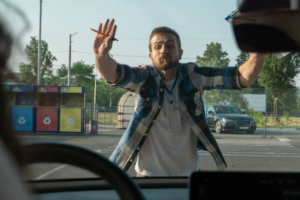 Młody instruktor stojący na placu treningowym w szkole jazdy na świeżym powietrzu, pokazujący gest zatrzymania rękami. Egzamin na prawo jazdy, kursy dla kierowców, koncepcja egzaminu - Zdjęcie, obraz