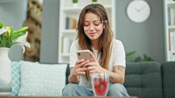 Νέα όμορφη ισπανόφωνη γυναίκα χρησιμοποιώντας smartphone κάθεται στον καναπέ χαμογελώντας στο σπίτι - Πλάνα, βίντεο