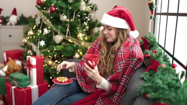 Giovane bella donna ispanica che fa colazione seduta sul divano vicino all'albero di Natale a casa - Filmati, video